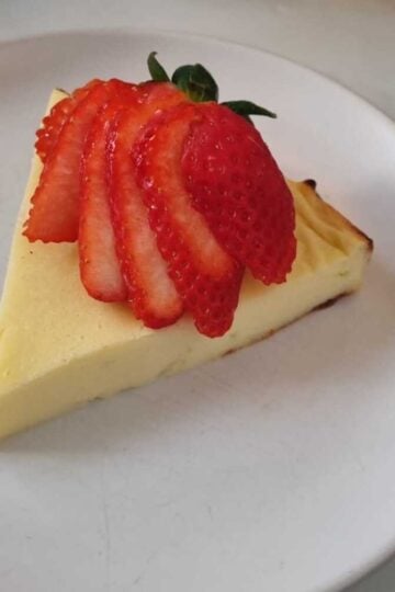 A slice of cottage cheese cheesecake garnished with a sliced strawberry on a white plate.