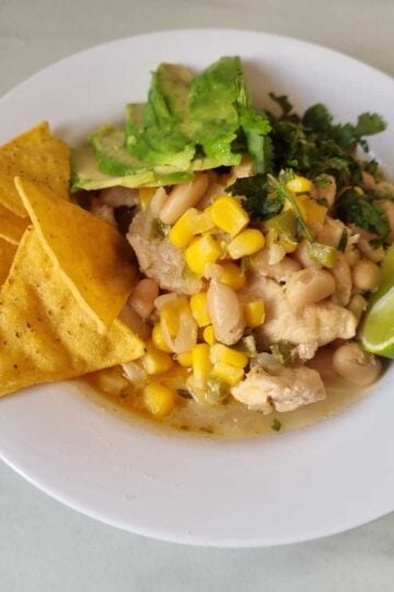 Bowl of white bean chicken chili garnished with a wedge of lime, tortilla chips, chopped fresh cilantro and sliced avocados.
