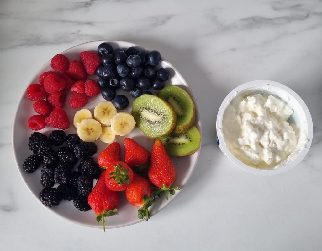 A packet of cottage cheese beside a plate of assorted fresh fruits, ready to be assembled into a high-protein cottage cheese with fruit breakfast bowl.